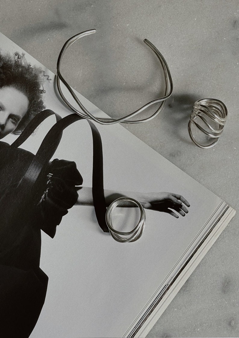 A stylized black and white photo featuring a hand with silver rings and a bracelet, laying over an open magazine with artistic imagery.