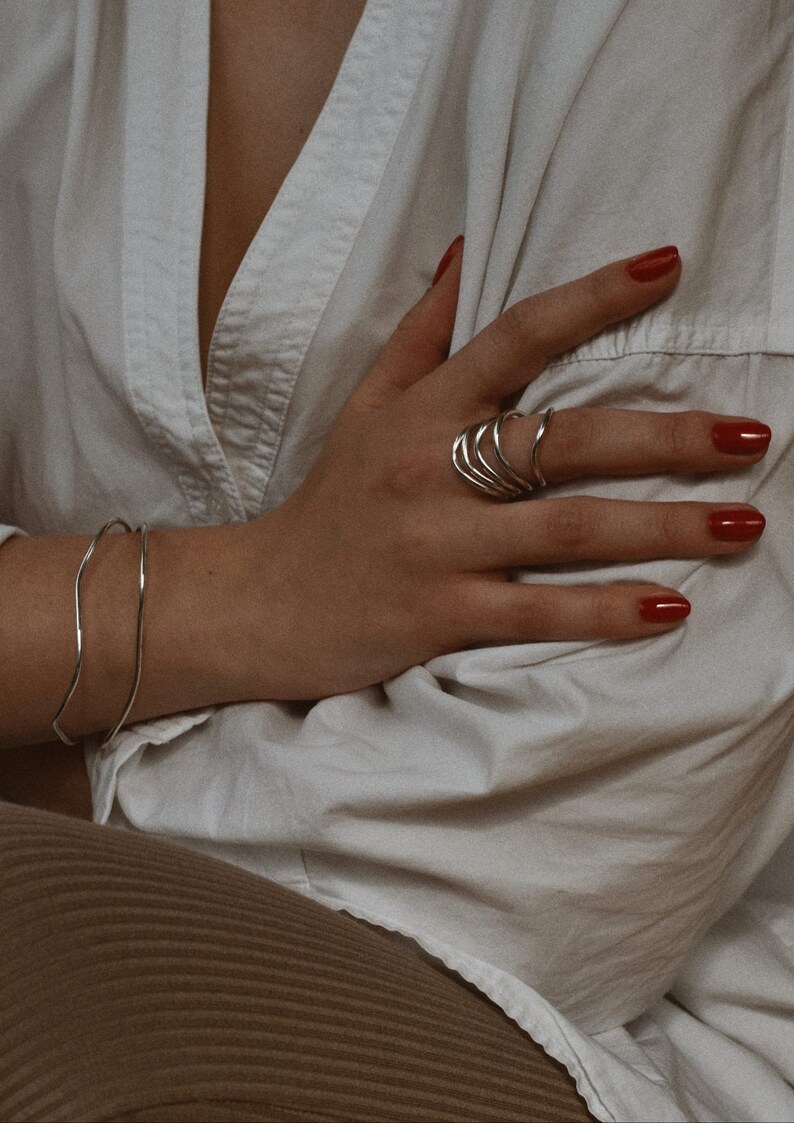 Hand with red nail polish showing silver spiral rings, resting on a white cloth with a relaxed, elegant posture.