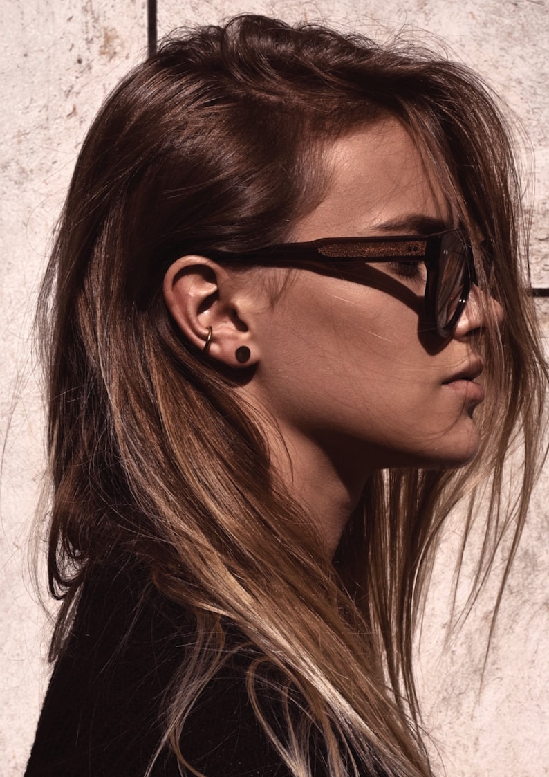 May include: A woman with long brown hair wearing black sunglasses and small gold hoop earrings. She is wearing a black shirt and has a dark brown stud earring in her left ear.
