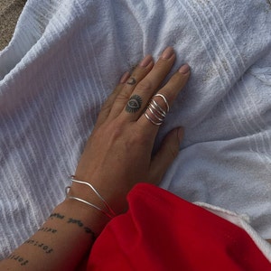 Hand resting on a white, fringed cloth over the sand, adorned with multiple silver rings, with visible arm tattoos, beside a swatch of red fabric.