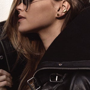 May include: A woman wearing a black leather jacket with a zipper and a gold ear cuff. She is also wearing a gold stud earring and sunglasses.