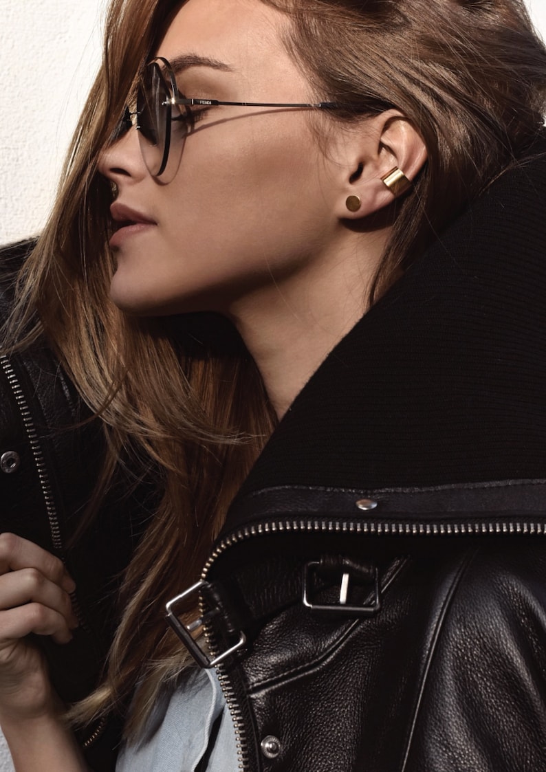 May include: A woman wearing a black leather jacket with a zipper and a gold ear cuff. She is also wearing a gold stud earring and sunglasses.
