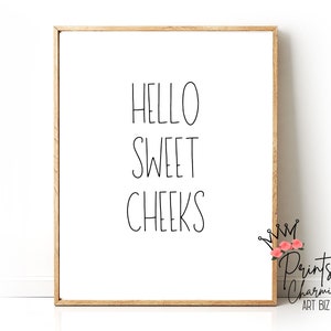 May include: A framed print with the words "HELLO SWEET CHEEKS" in black script on a white background. The frame is a light brown color. The lower right corner has a logo with text and floral accents.