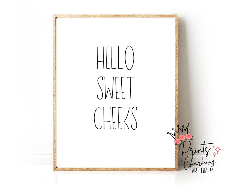 May include: A framed print with the words "HELLO SWEET CHEEKS" in black script on a white background. The frame is a light brown color. The lower right corner has a logo with text and floral accents.