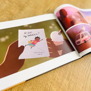 May include: A hand holds a card with a drawing of a superhero child flying. The card says "For Dad, my SUPERHERO" and "Super Hamira, aged 72".