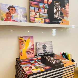 May include: A stack of 12 black and orange hardback books with the title "The Fell We Climb" on the top book. The book cover features a collage of colourful illustrations. There is a smaller book with the same title and a bookmark with a cartoon character on it.