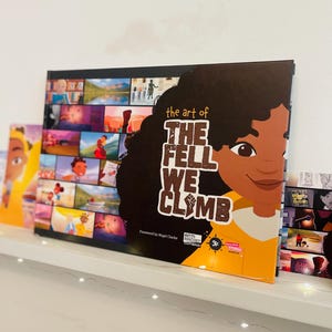 May include: A yellow and brown book titled "The Art of The Fell We Climb" with a colourful collage of images on the cover. The book features a young girl with brown skin and curly hair. The book is on a white shelf with a string of white lights.