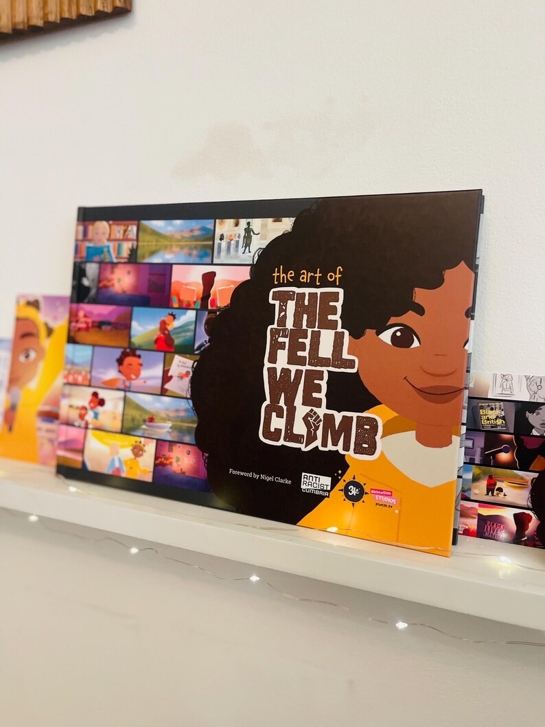 May include: A yellow and brown book titled "The Art of The Fell We Climb" with a colourful collage of images on the cover. The book features a young girl with brown skin and curly hair. The book is on a white shelf with a string of white lights.