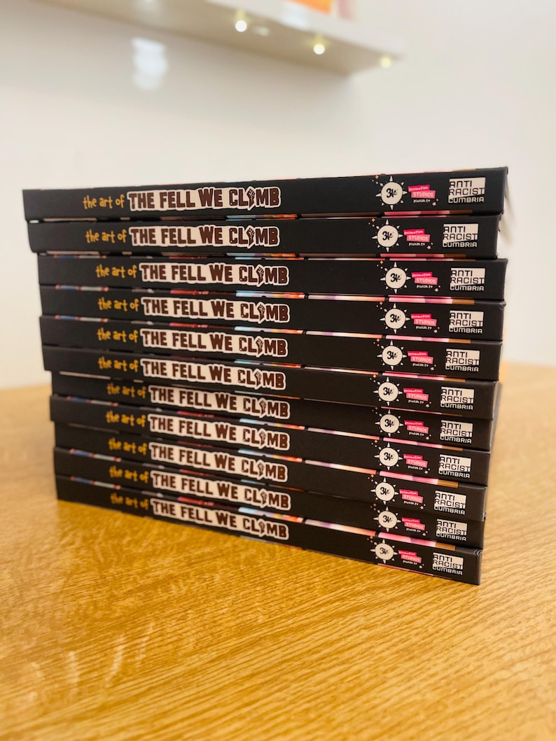 May include: A stack of 10 black books with the title "The Fell We Climb" and the words "the art of" printed on each book. Each book has a sticker on the spine that says "Anti-Racist Cumbria" and a number "34" printed on the sticker.