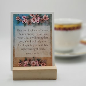 May include: A white card with a floral design and a bible verse from Isaiah 41:10. The verse reads "Fear not, for I am with you; Be not dismayed, for I am your God. I will strengthen you, Yes, I will help you, I will uphold you with My righteous right hand. ISAIAH 41:10". The card is sitting on a wooden stand.