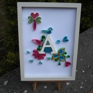 Può includere: Immagine incorniciata in bianco con una lettera A bianca circondata da fiori di carta colorati e un uccello blu. I fiori sono rosa, verdi, blu e gialli.