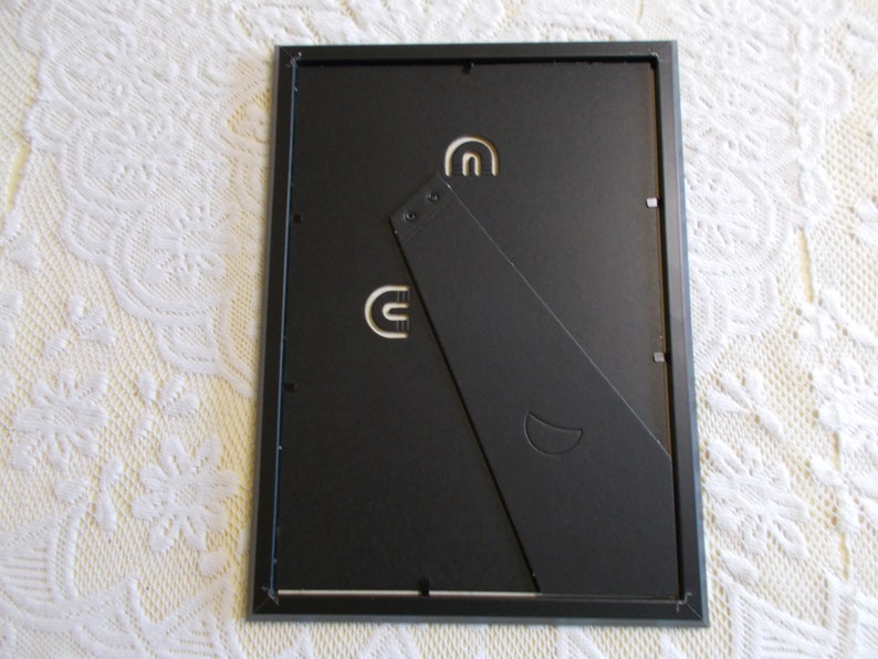 May include: The back of a black picture frame with a fold-out easel stand. The frame has a silver border and a crescent moon logo.