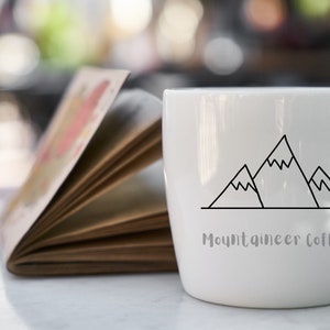 Op de afbeelding: Een witte keramische koffiemok met een zwarte omtrek van drie bergen en de tekst "Mountaineer Coffee".