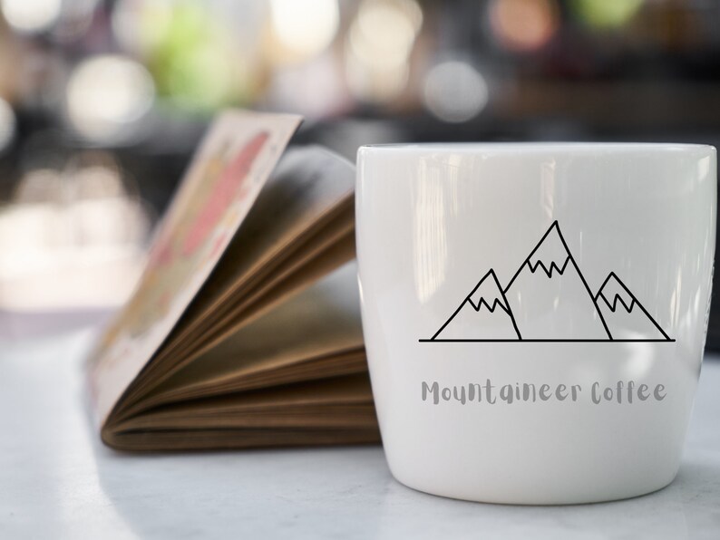 Op de afbeelding: Een witte keramische koffiemok met een zwarte omtrek van drie bergen en de tekst "Mountaineer Coffee".