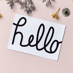 Op de afbeelding: Een witte kaart met het woord "hello" geschreven in zwarte cursieve letters. De kaart is omringd door kleine decoratieve items, waaronder gedroogde bloemen, kralen en metalen knopen.