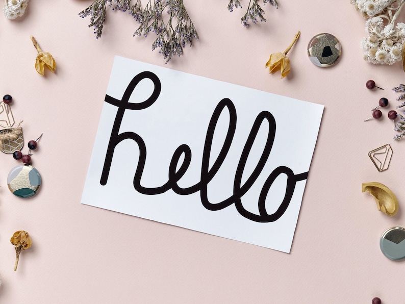 Op de afbeelding: Een witte kaart met het woord "hello" geschreven in zwarte cursieve letters. De kaart is omringd door kleine decoratieve items, waaronder gedroogde bloemen, kralen en metalen knopen.