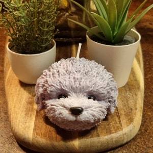 May include: A lavender-coloured candle shaped like a fluffy dog's head. The candle is sitting on a wooden chopping board.