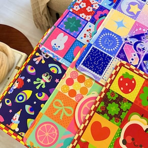 Può includere: Una collezione di oggetti piatti rettangolari con vivaci motivi a griglia multicolore. I disegni includono un coniglio bianco, una rana verde, occhi colorati, un clown, agrumi, un cuore rosso, una fragola e motivi celestiali in varie tonalità.