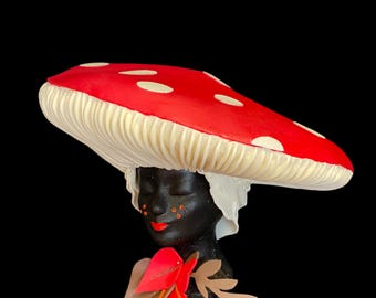 May include: A black ceramic bust with a red and white mushroom hat. The bust has a red mouth and orange dots on the face. The bust is wearing a red and orange flower.