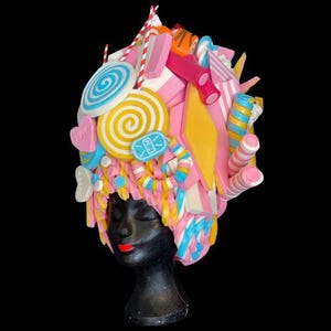 Pode incluir: Um adorno de cabeça extravagante com tema de doces, apresentando uma variedade de doces coloridos. O design inclui pirulitos, bastões de doces e outras guloseimas em tons de rosa, amarelo, azul e branco. O adorno de cabeça é exibido em uma cabeça de manequim preta.