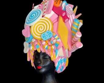 May include: A whimsical candy-themed headpiece featuring an assortment of colorful sweets. The design includes lollipops, candy sticks, and various other treats in shades of pink, yellow, blue, and white. The headpiece is displayed on a black mannequin head.