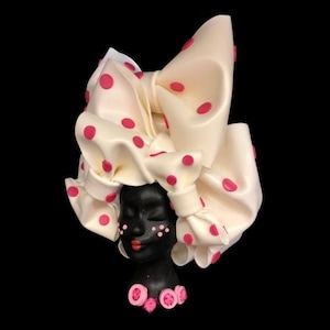 May include: A white ceramic bust with a pink polka dot head wrap. The bust has a black face with pink accents and closed eyes. The bust is wearing a pink flower necklace.