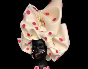 May include: A white ceramic bust with a pink polka dot head wrap. The bust has a black face with pink accents and closed eyes. The bust is wearing a pink flower necklace.