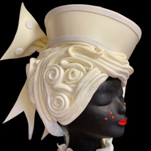 May include: Ivory-colored headpiece with a decorative design. The headpiece features a structured brim, a bow with circular accents, and swirling, textured details. The headpiece is displayed on a black mannequin head with red lips and orange accents.