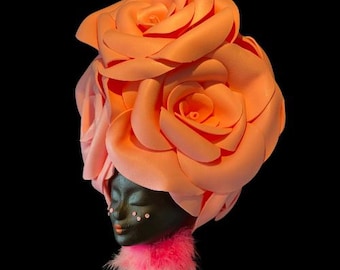 May include: A black face with pink and peach fabric roses forming a crown on top of the head. The roses are made of fabric and are in a variety of sizes. The face is painted with black paint and has a pink and white feather boa around the neck.