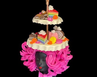 May include: A pink wig with a white tiered cake stand topped with colorful cakes, cookies, and candies. The stand is on top of the wig.