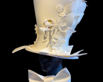 May include: A white tall hat with a wide brim, decorated with white flowers and a small white bird. The hat is on a black mannequin wearing a white bow.