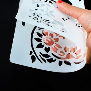 May include: White plastic stencil with a floral design. The stencil is folded in half, showing the design on both sides.