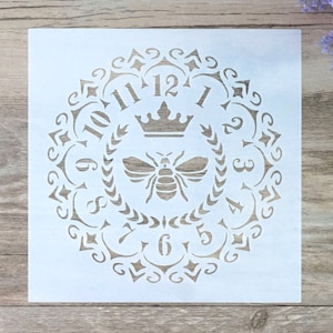 Può includere: Stencil bianco con un design decorativo che presenta un'ape, una corona e numeri romani attorno al bordo. Lo stencil è per l'artigianato e la decorazione.