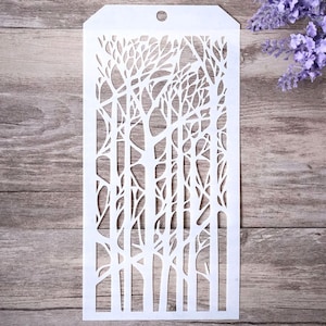 Puede incluir: Plantilla de plástico blanco con un diseño de un bosque de árboles con ramas desnudas. La plantilla es rectangular y tiene un agujero en la parte superior para colgar.