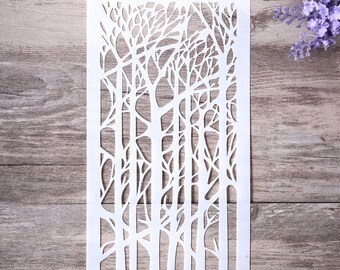 May include: White plastic stencil with a design of a forest of trees with bare branches. The stencil is rectangular and has a hole in the top for hanging.