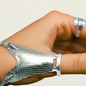 May include: A silver finger splint and hand brace. The brace is made of a flexible, mesh-like material and is connected to a ring on the thumb and a ring on the index finger. The ring on the index finger is a different design.