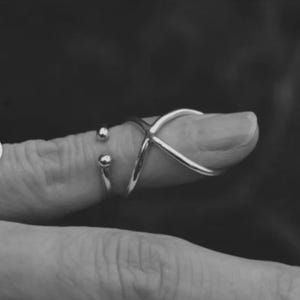 May include: A silver-toned ring with an X-shaped design and two small ball accents. The ring is worn on a finger, with the design crossing over the top of the finger. The background is blurred.