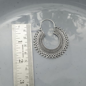 Könnte beinhalten: Silberner Creolen-Ohrring mit einem aufwendigen, kreisförmigen Design. Der Ohrring hat eine Reihe kleiner, erhabener Punkte am äußeren Rand und in den inneren Kreisen. Ein Lineal liegt daneben.