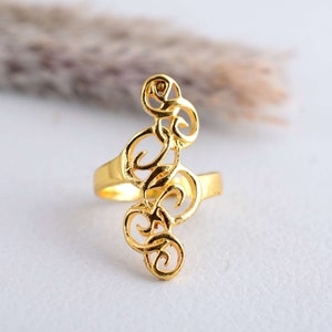 Könnte beinhalten: Ein goldfarbener Ring mit einem durchbrochenen Design aus wirbelnden, abstrakten Formen. Der Ring hat einen Band und ein längliches dekoratives Element, das sich nach oben erstreckt. Der Ring ist vor einem weichen, neutralen Hintergrund platziert.