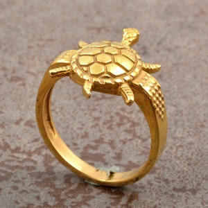 Op de afbeelding: Een goudkleurige ring met een gedetailleerd schildpadontwerp. De schild en ledematen van de schildpad zijn ingewikkeld getextureerd en de ringband is glad. De ring staat tegen een neutrale achtergrond, wat de gouden kleur en het vakmanschap benadrukt.