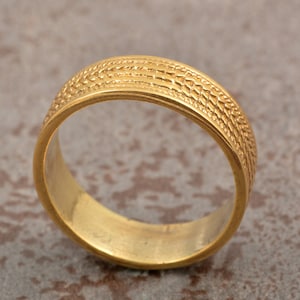May include: A gold ring with a textured band. The ring's surface has a repeating pattern of small, raised lines and shapes, creating a detailed design. The ring is a simple band style, suitable for everyday wear. The ring is made of gold.