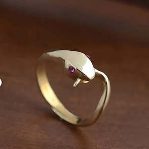 May include: A gold-colored ring in the shape of a snake, with two small, round, pink gemstones for eyes. The snake's head is slightly raised, and the ring is set against a brown background.