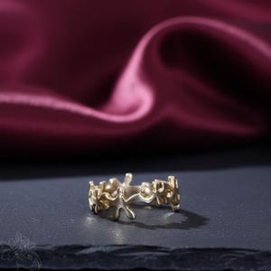 May include: A gold-colored ring with a unique design featuring small, abstract human figures. The ring is set against a dark surface with a blurred, burgundy fabric in the background. The ring's design is intricate and detailed.
