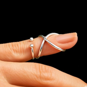 Könnte beinhalten: Ein silberner Fingerring mit einem einzigartigen Design. Der Ring hat zwei kleine, runde Akzente und ein Kreuzdesign, das sich um den Finger wickelt. Der Ring wird auf einem Finger vor schwarzem Hintergrund gezeigt.