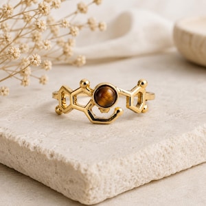May include: A gold-toned ring featuring a molecular structure design with a central circular brown tiger's eye gemstone. The metal band is shaped into interconnected hexagonal and pentagonal geometric forms, creating a scientific aesthetic.