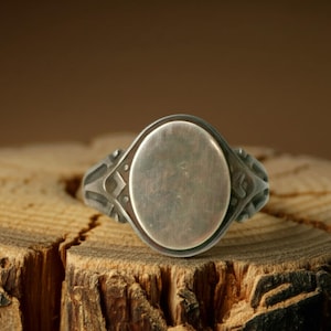 May include: A silver signet ring with an oval face. The ring has decorative detailing on the shoulders and a slightly oxidized finish. The ring is displayed on a wooden surface.