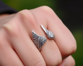 May include: A silver-colored ring with a wing design. The ring features two detailed wings, one on each side, creating an open design. The ring is worn on a finger, with a blurred green background.