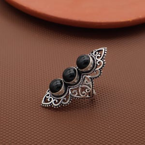 May include: A silver-colored ring featuring three round, black gemstones set in a row. The ring has an ornate, filigree design with intricate patterns and a decorative, elongated shape.