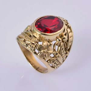 Puede incluir: Anillo dorado con una gema roja ovalada y facetada. El anillo presenta un diseño ornamentado con motivos florales y de mariposas. La banda y el engaste son de color oro cálido, creando un contraste llamativo con la piedra roja.