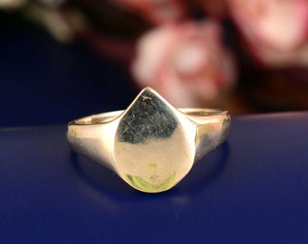 May include: A gold signet ring featuring a smooth, polished surface with a distinct teardrop or pear-shaped face. The band is solid and tapers toward the back, showcasing a reflective metallic finish throughout the entire piece of jewelry.